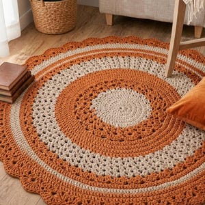 May include: A round, hand-crocheted rug in shades of orange and cream. The rug features a scalloped edge and concentric circles of color and openwork. The rug is placed on a wooden floor, with a plant and furniture in the background.