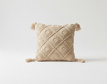 May include: A square, beige crochet pillow with a floral pattern and diamond grid design. The pillow has four tassels, one at each corner. The crochet work is detailed and the pillow is a neutral color.