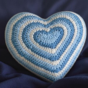 May include: A handmade, heart-shaped pillow with concentric circles of light blue and white crochet stitches. The pillow is set against a dark blue background, creating a soft, textured appearance. This decorative item is perfect for adding a touch of handmade charm.