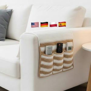 Pode incluir: Um organizador de apoio de braço de sofá de crochê em branco e bege. O organizador tem três bolsos, cada um com um controle remoto. O design apresenta um padrão listrado nos bolsos e uma seção superior branca lisa. Ele está sobre o braço de um sofá branco.