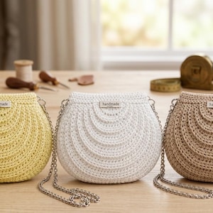 Peut inclure: Trois sacs à main crochetés faits à la main en jaune, blanc et beige, chacun avec une bandoulière en chaîne argentée. Les sacs ont une forme arrondie avec un motif ondulé décoratif. Chaque sac porte une petite étiquette « fait main ».