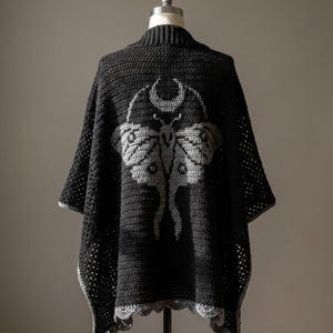 May include: A black crochet poncho featuring a large gray moth graphic on the back, topped with a crescent moon design. The garment has a ribbed collar, a textured stitch pattern throughout, and a scalloped gray border along the bottom edge.