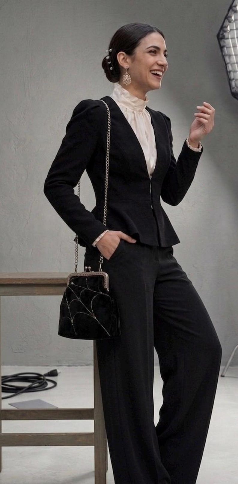 May include: Black peplum blazer and matching trousers. The blazer has a fitted silhouette with a chain strap bag. The bag is black with a silver spiderweb design. The outfit is completed with a white blouse.