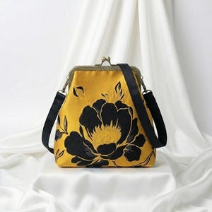 May include: Mustard yellow handbag with a black floral design. The bag has a kiss-lock closure, a black shoulder strap, and a gold-colored frame. The floral pattern is a large, stylized peony.