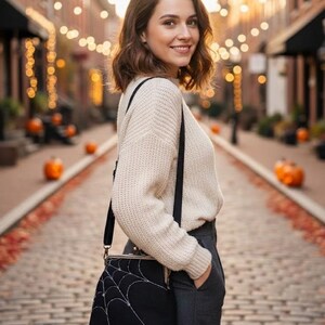 May include: A black crossbody bag with a white spiderweb design. The bag has a clasp closure and a long black strap. The person is wearing a cream-colored sweater and gray pants. The background shows a brick-paved street with pumpkins and string lights.