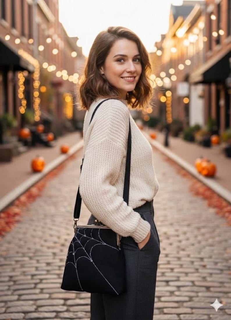 May include: A black crossbody bag with a white spiderweb design. The bag has a clasp closure and a long black strap. The person is wearing a cream-colored sweater and gray pants. The background shows a brick-paved street with pumpkins and string lights.