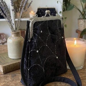 May include: Black velvet crossbody bag with a silver clasp and strap. The bag features a spiderweb design embroidered with silver thread and embellished with small, sparkling rhinestones. A candle and vase of lavender are in the background.