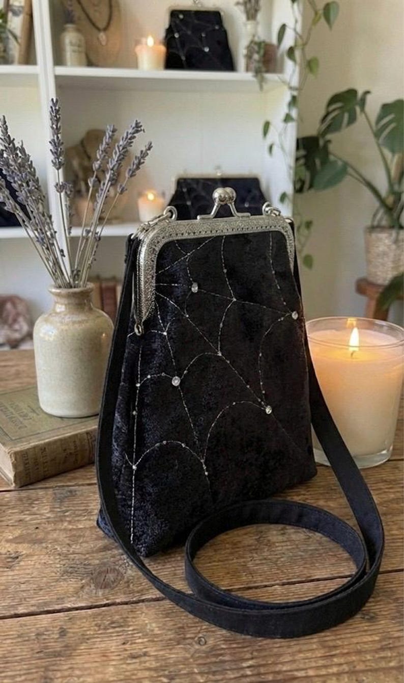 May include: Black velvet crossbody bag with a silver clasp and strap. The bag features a spiderweb design embroidered with silver thread and embellished with small, sparkling rhinestones. A candle and vase of lavender are in the background.