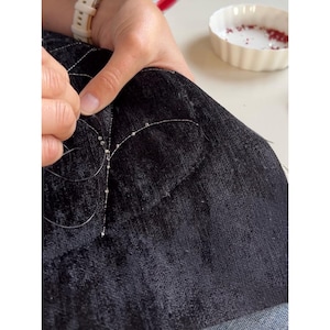 May include: A close-up of a person's hands sewing beads onto a dark blue fabric. The beads are small and silver, and the thread is barely visible. A small white bowl with red beads is in the background.