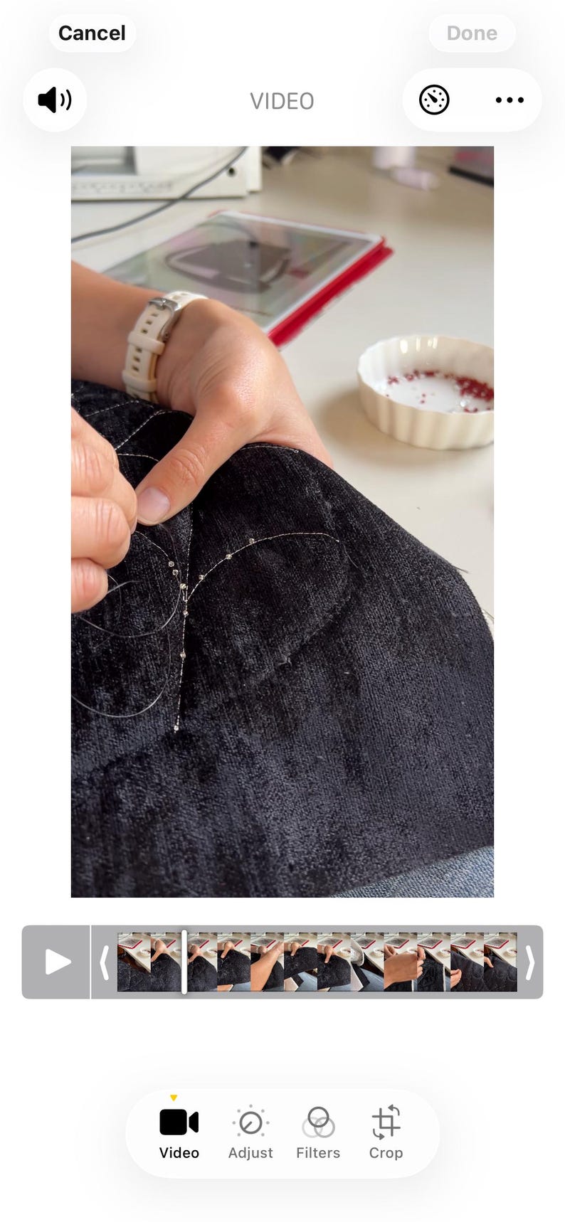 May include: A close-up of a person's hands sewing beads onto a dark blue fabric. The beads are small and silver, and the thread is barely visible. A small white bowl with red beads is in the background.
