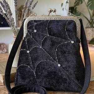May include: Black velvet crossbody bag with a silver clasp and strap. The bag features a spiderweb design embroidered with silver thread and embellished with small, sparkling gems. A stylish accessory for everyday use.