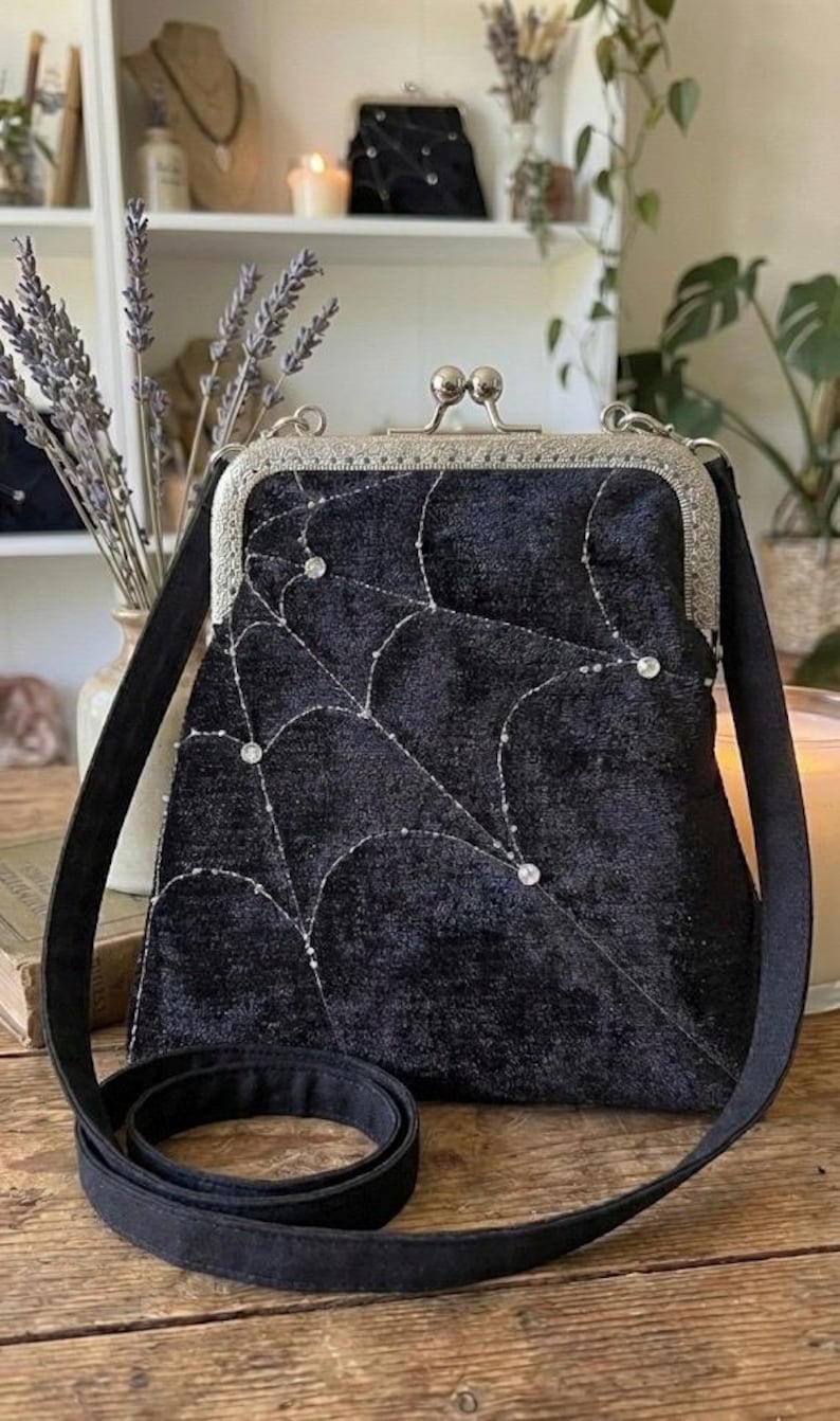 May include: Black velvet crossbody bag with a silver clasp and strap. The bag features a spiderweb design embroidered with silver thread and embellished with small, sparkling gems. A stylish accessory for everyday use.