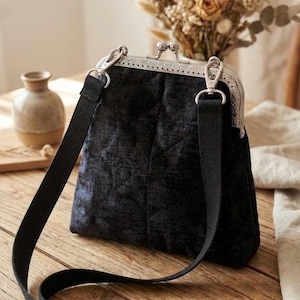 May include: A black velvet handbag with a silver clasp and a black shoulder strap. The bag has a quilted pattern and a rectangular shape. The background includes a vase, dried flowers, and a wooden table.