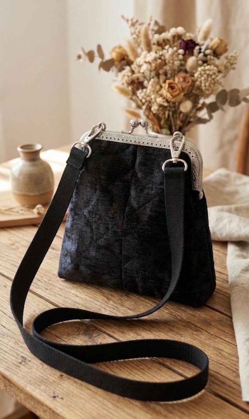 May include: A black velvet handbag with a silver clasp and a black shoulder strap. The bag has a quilted pattern and a rectangular shape. The background includes a vase, dried flowers, and a wooden table.