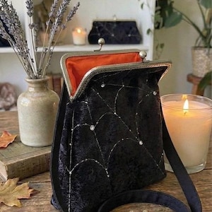 May include: Black velvet handbag with a silver clasp and a spiderweb design. The interior is orange. The bag has a black strap and is displayed on a wooden surface with acorns and fall leaves. A lit candle and a vase of lavender are in the background.