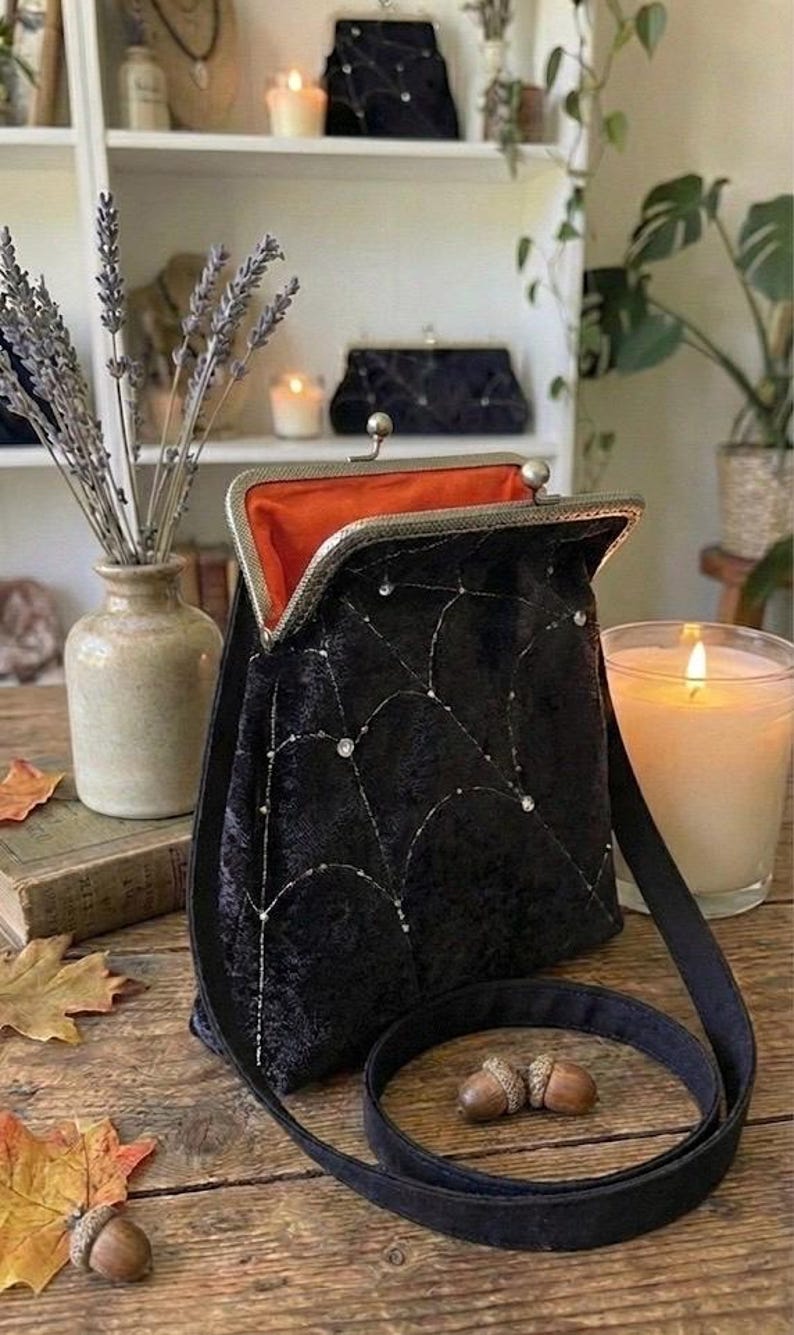 May include: Black velvet handbag with a silver clasp and a spiderweb design. The interior is orange. The bag has a black strap and is displayed on a wooden surface with acorns and fall leaves. A lit candle and a vase of lavender are in the background.