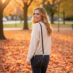 May include: A black crossbody bag with a white spiderweb design. The bag is worn over the shoulder. The person is wearing a cream-colored sweater and dark blue jeans. The background is a park with autumn leaves.