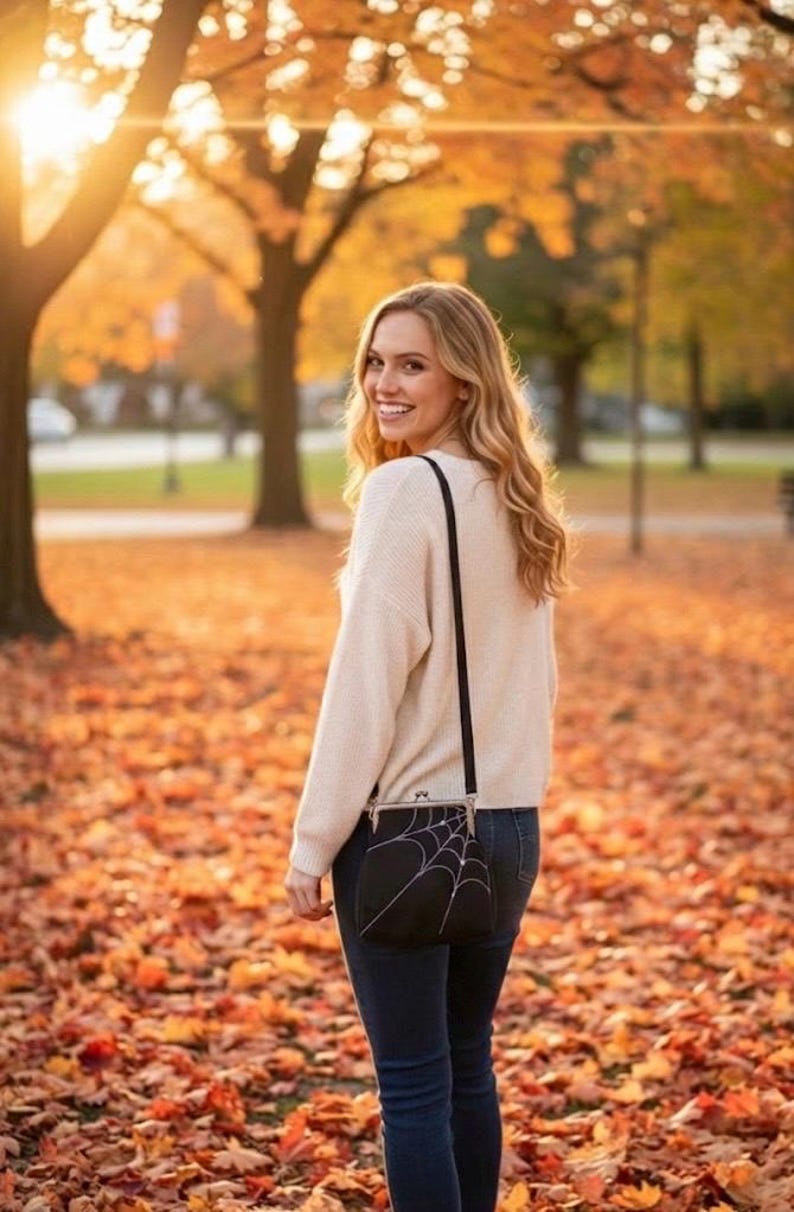 May include: A black crossbody bag with a white spiderweb design. The bag is worn over the shoulder. The person is wearing a cream-colored sweater and dark blue jeans. The background is a park with autumn leaves.