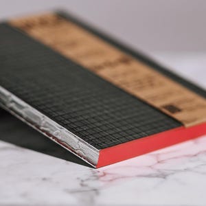 Pode incluir: Close-up de um caderno com capa preta texturizada e borda vermelha. A seção superior é marrom claro. As páginas são brancas e o caderno está sobre uma superfície de mármore.