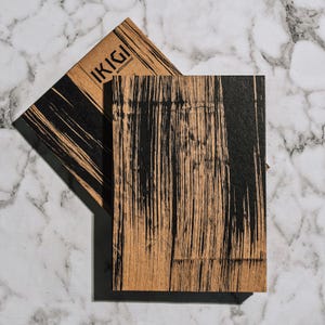 May include: Two rectangular notebooks with a wood grain texture and black brushstroke design. The top notebook is partially visible, displaying the text "IKIGI" in a bold font. The notebooks are set against a marble background.