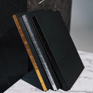 Könnte beinhalten: Ein Stapel von vier schwarzen Notizbüchern mit gold- und silberfarbenen Kanten. Die Notizbücher lehnen an einem schwarzen Marmorblock auf einer weißen Marmoroberfläche. Der goldene Rand befindet sich links, gefolgt von silbernen und schwarzen Rändern.