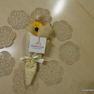 Può includere: Un bouquet di fiori gialli avvolto in carta beige, legato con un nastro e circondato da sei centrini all'uncinetto beige. Un'etichetta sul bouquet recita "Homemade by One Hook."