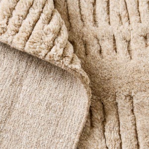 Puede incluir: Primer plano de una manta beige texturizada. La manta tiene una superficie suave y afelpada con un patr&oacute;n acanalado. La parte inferior de la manta es de un tono beige m&aacute;s claro y liso. Los bordes est&aacute;n cuidadosamente acabados.
