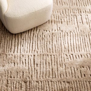 Puede incluir: Una alfombra beige texturizada con un patr&oacute;n lineal vertical. La alfombra est&aacute; hecha de un material suave y tiene una apariencia afelpada. Una parte de un sill&oacute;n color crema es visible en la esquina superior izquierda.