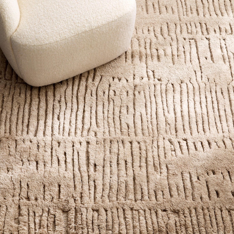 Puede incluir: Una alfombra beige texturizada con un patr&oacute;n lineal vertical. La alfombra est&aacute; hecha de un material suave y tiene una apariencia afelpada. Una parte de un sill&oacute;n color crema es visible en la esquina superior izquierda.