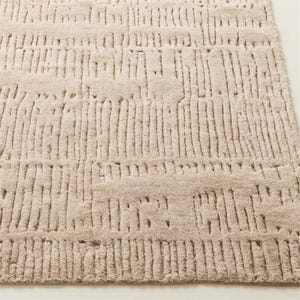 Puede incluir: Una alfombra rectangular beige con un patr&oacute;n texturizado en relieve. El dise&ntilde;o presenta l&iacute;neas verticales y horizontales, creando una apariencia tejida. La alfombra est&aacute; hecha de un material suave, adecuado para una sala de estar o un dormitorio.