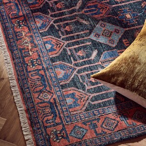 K&ouml;nnte beinhalten: Nahaufnahme eines gemusterten Teppichs mit dunkelblauem Hintergrund und roten und blauen geometrischen Designs. Der Teppich hat einen Fransenrand und ein goldfarbenes Kissen in der Ecke.