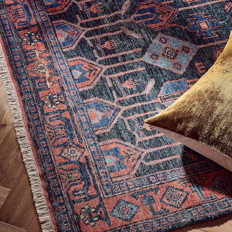 K&ouml;nnte beinhalten: Nahaufnahme eines gemusterten Teppichs mit dunkelblauem Hintergrund und roten und blauen geometrischen Designs. Der Teppich hat einen Fransenrand und ein goldfarbenes Kissen in der Ecke.