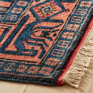 K&ouml;nnte beinhalten: Nahaufnahme eines gewebten Teppichs mit einem geometrischen Muster in Blau- und Korallt&ouml;nen. Der Teppich hat einen Fransenrand und liegt auf einem Holzboden. Das Design zeigt Diamanten- und Zickzackformen.
