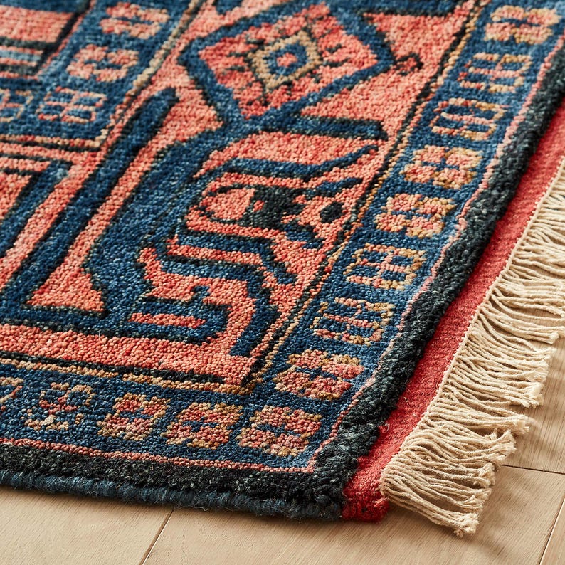 K&ouml;nnte beinhalten: Nahaufnahme eines gewebten Teppichs mit einem geometrischen Muster in Blau- und Korallt&ouml;nen. Der Teppich hat einen Fransenrand und liegt auf einem Holzboden. Das Design zeigt Diamanten- und Zickzackformen.
