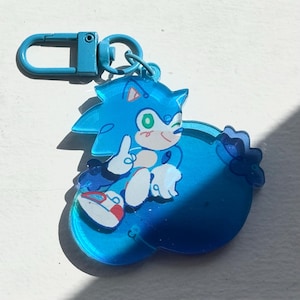 May include: A blue acrylic keychain featuring the character Sonic the Hedgehog. The keychain has a blue clasp and a charm depicting Sonic winking and giving a thumbs up. The charm is a light blue color with white and red accents.