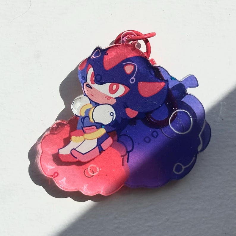 May include: A colorful acrylic charm featuring a cartoon character with red and blue accents. The character is sitting on a pink cloud-shaped base. The charm has a red loop for attachment.