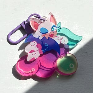 May include: A colorful acrylic keychain featuring a cartoon character with white fur, blue eyes, and pink accents. The character is on a cloud-shaped base with pink, purple, and green gradients. A purple clasp is attached for easy use.