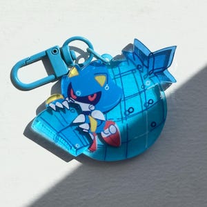 May include: A blue Metal Sonic keychain with a matching clasp. The keychain features a cartoon character with a blue body, red eyes, and yellow accents. The character is on a blue, globe-shaped charm.