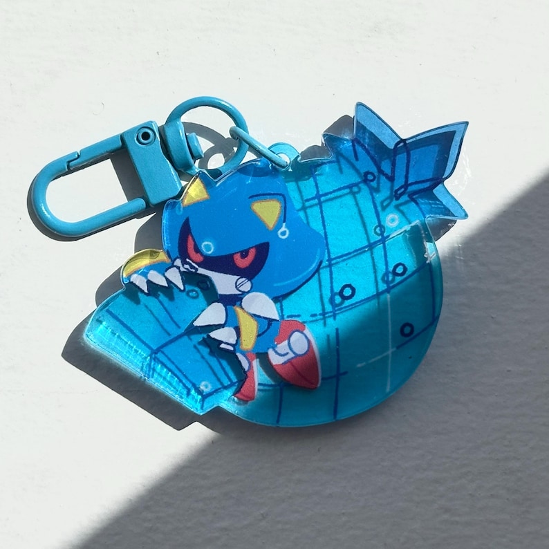 May include: A blue Metal Sonic keychain with a matching clasp. The keychain features a cartoon character with a blue body, red eyes, and yellow accents. The character is on a blue, globe-shaped charm.