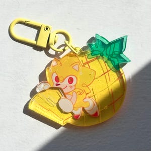 May include: A yellow acrylic keychain featuring a cartoon character with yellow fur and red eyes, perched on a yellow pineapple. The keychain has a yellow clasp and a green leaf accent on the pineapple.