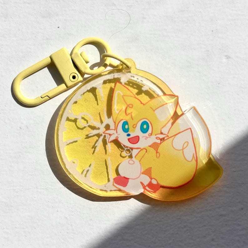 May include: A yellow keychain featuring a cartoon fox character with blue eyes and a red and white outfit. The character is set against a yellow lemon slice design. The keychain has a yellow clasp.