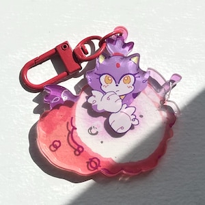 May include: A clear acrylic keychain featuring a purple cartoon character with a crown and orange eyes. The character is set against a pink and white background with a red lobster claw clasp.