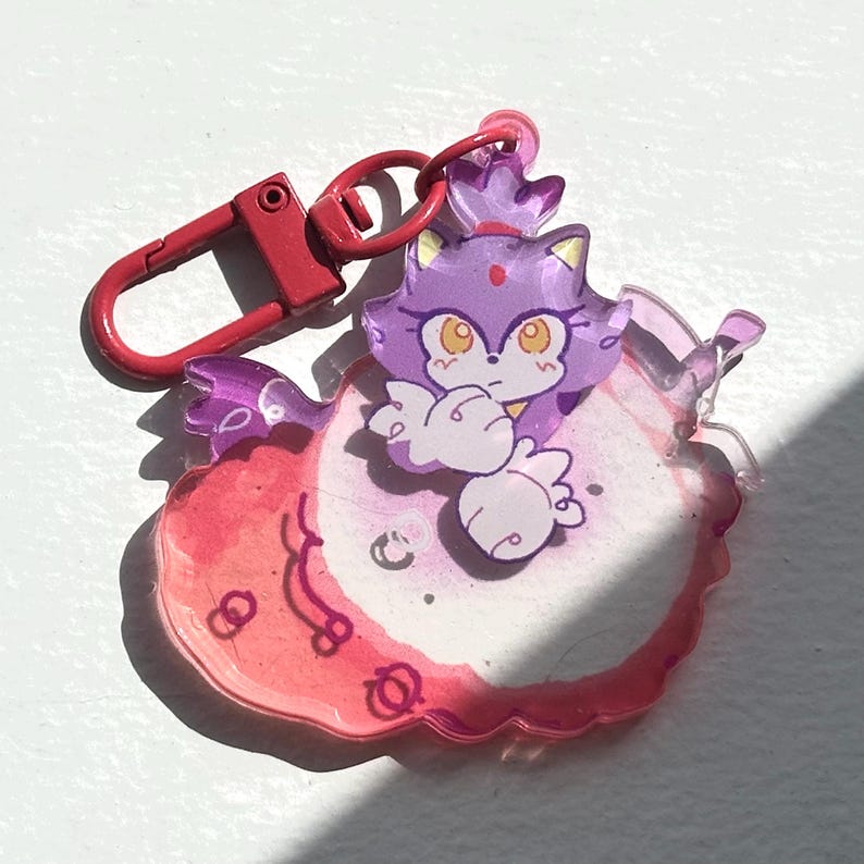 May include: A clear acrylic keychain featuring a purple cartoon character with a crown and orange eyes. The character is set against a pink and white background with a red lobster claw clasp.