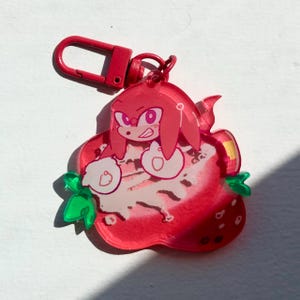 May include: A red acrylic keychain featuring a cartoon character, Knuckles, from the Sonic the Hedgehog series, inside a strawberry. The keychain has a red clasp and green leaves. The strawberry is red with white and pink details.