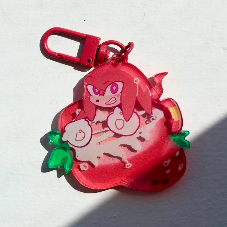 May include: A red acrylic keychain featuring a cartoon character, Knuckles, from the Sonic the Hedgehog series, inside a strawberry. The keychain has a red clasp and green leaves. The strawberry is red with white and pink details.