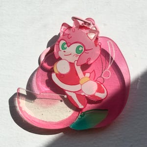 May include: A pink acrylic charm featuring a cartoon character with pink hair, green eyes, and a red dress. The charm has a pink loop for attachment and is set against a white background. The character is in a dynamic pose.