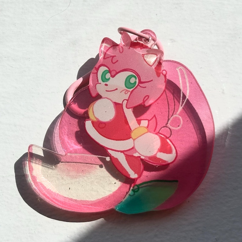 May include: A pink acrylic charm featuring a cartoon character with pink hair, green eyes, and a red dress. The charm has a pink loop for attachment and is set against a white background. The character is in a dynamic pose.