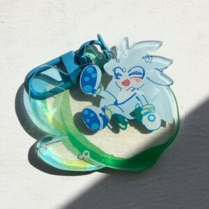 May include: A clear acrylic charm featuring a cartoon character with spiky hair, blue shoes, and a red mouth. The charm is attached to a blue clasp and a translucent, green-tinted base. The design is outlined in black.