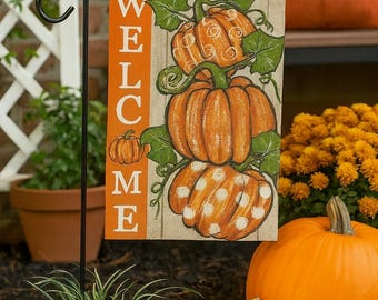 May include: A fall-themed garden flag with the word "WELCOME" in orange. The flag features three orange pumpkins with green leaves. Several pumpkins of different sizes and colors are in front of the flag.