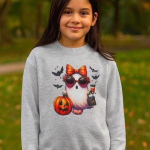 May include: A light grey crewneck sweatshirt featuring a graphic print of a white ghost wearing a large orange bow and round sunglasses. The ghost holds a drink and a black bag next to a carved orange jack-o'-lantern, surrounded by black bats.
